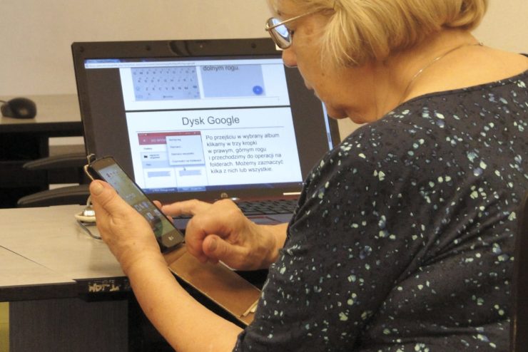 Seniorka korzystająca ze smartfona. W tle znajduje się laptop, na którym wyswietla się prezentacja dotyczaca Dysku Google.