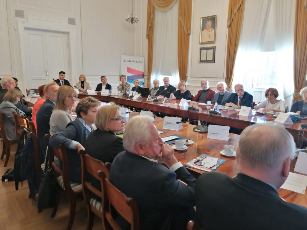 Zdjęcie ilustrujące posiedzenie Małopolskiej Rady ds. Polityki Senioralnej w dniu 5 grudnia 2022 r. Przy stole siedzą członkowie Rady. W tle rolap Małopolski.