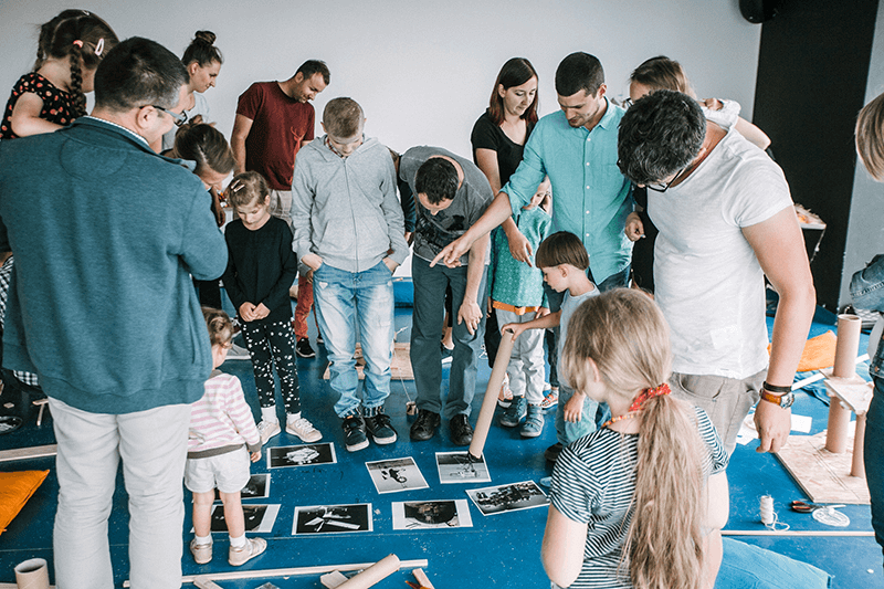 Zdjęcie ilustrujące zajęcia międzypokoleniowe. Grupa osób w różnym wieku pochyla się nad rozrzuconymi na podłodze fotografiami. Wskazują palcami zdjęcia.