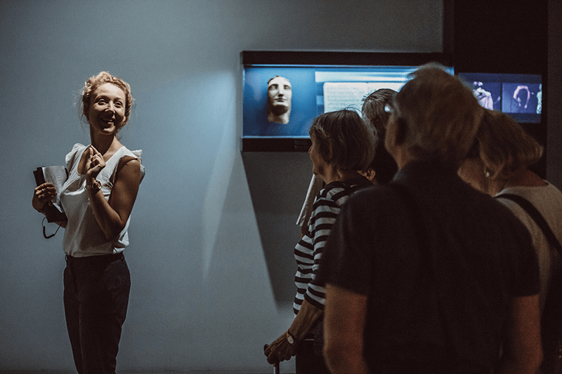 Zdjęcie ilustrujące zwiedzanie wystawy w Cricotece, na zdjęciu uśmiechnięta młoda kobieta z książką w ręce, której przygląda się grupa seniorek. W tle element wystawy.