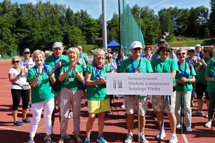 Seniorzy ze Stowarzyszenia Wielicki Uniwersytet Trzeciego Wieku stoją na boisku z zielonych koszulkach i pozują do zdjęcia z tabliczką z nazwą Stowarzyszenia